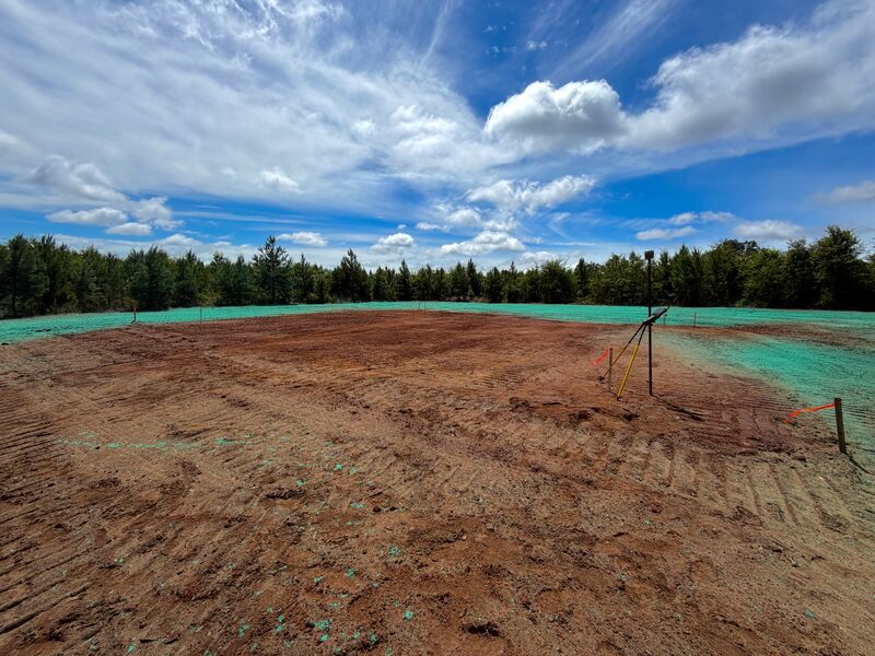 Graded red-clay house pad freshly hydroseeded with bright green mulch under a blue sky with clouds, ready for staking